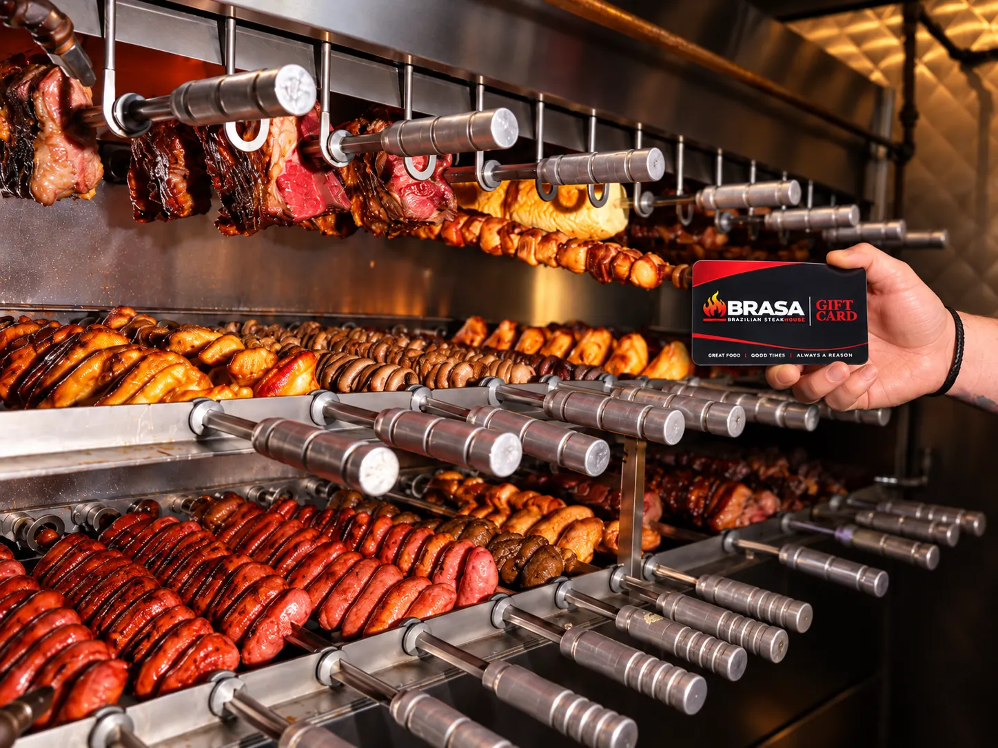 Brasa Gift Card held in front of Brazilian churrasco skewers on the grill